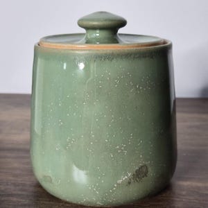 May include: A green ceramic jar with a lid. The jar has a speckled finish and a rounded shape. The lid is a darker green with a small knob on top. The jar sits on a wooden surface.