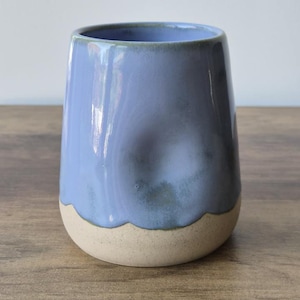 May include: A ceramic tumbler with a smooth, periwinkle blue glazed upper section and a textured, speckled beige base. The vessel features a distinct thumb-shaped indentation on one side and a rounded, handleless silhouette with a wide opening.
