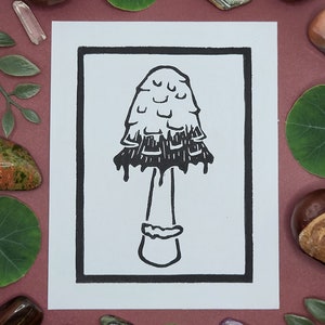 May include: A black and white line drawing of a mushroom with a dripping cap, printed on a white card with a black border. The card is on a burgundy background.