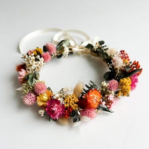May include: A floral crown made with dried flowers in shades of pink, orange, and yellow. The crown is made with a variety of flowers, including roses, daisies, and wildflowers.