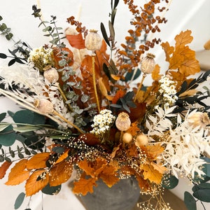 K&ouml;nnte beinhalten: Eine getrocknete Blumenarrangement in einer grauen Keramikvase. Die Anordnung enth&auml;lt getrockneten Eukalyptus, braune und orangefarbene Bl&auml;tter, wei&szlig;e getrocknete Blumen und beige Samenkapseln.