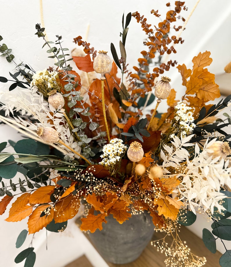 K&ouml;nnte beinhalten: Eine getrocknete Blumenarrangement in einer grauen Keramikvase. Die Anordnung enth&auml;lt getrockneten Eukalyptus, braune und orangefarbene Bl&auml;tter, wei&szlig;e getrocknete Blumen und beige Samenkapseln.