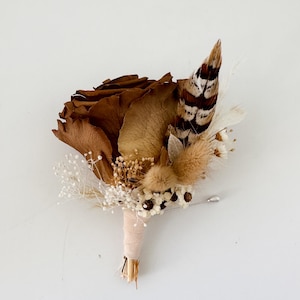 May include: A rustic boutonniere featuring a large, dried brown rose, accented with a patterned feather, small white flowers, and tan dried grasses. The stems are wrapped in a light beige fabric, secured with a silver pin.