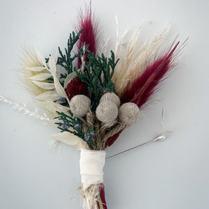 May include: A small bouquet of dried flowers with a white ribbon and a silver pin. The bouquet includes burgundy, white, and beige dried grasses, green foliage, and small beige pom-poms.