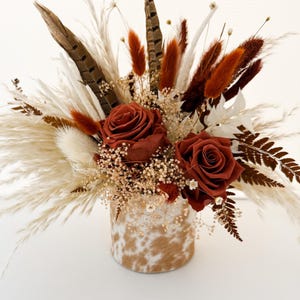 May include: A floral arrangement featuring two large rust-colored roses, dried white pampas grass, brown feathers, and small white baby's breath flowers. The bouquet is contained in a cylindrical vase with a tan and white cowhide-style pattern.
