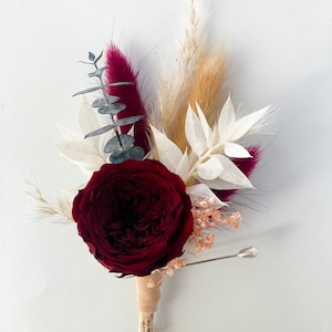May include: A burgundy rose with dried pampas grass, white fluffy dried flowers, and eucalyptus leaves. The bouquet is tied with a beige ribbon and secured with a pin.