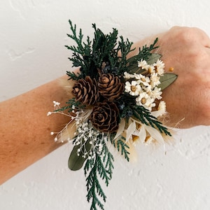 May include: A wrist corsage featuring a mix of dried flowers and greenery. The arrangement includes brown pinecones, white flowers, and dark green foliage. The corsage is accented with small white sprigs and a few dark blue berries.
