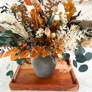 K&ouml;nnte beinhalten: Eine Trockenblumen-Arrangement in einer grauen Keramikvase. Die Anordnung enth&auml;lt getrockneten Eukalyptus, Pampasgras und braune Bl&auml;tter. Die Vase steht auf einem Holztablett.