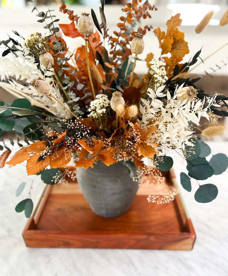 K&ouml;nnte beinhalten: Eine Trockenblumen-Arrangement in einer grauen Keramikvase. Die Anordnung enth&auml;lt getrockneten Eukalyptus, Pampasgras und braune Bl&auml;tter. Die Vase steht auf einem Holztablett.