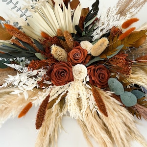 May include: A dried floral arrangement with a mix of pampas grass, dried roses, and other dried foliage in shades of brown, white, and green.