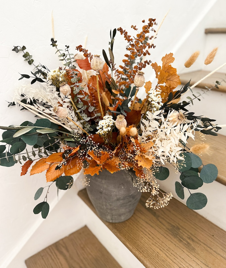 K&ouml;nnte beinhalten: Ein Trockenblumenarrangement in einer grauen Keramikvase. Die Anordnung enth&auml;lt wei&szlig;e, braune und orangefarbene Trockenblumen und Gr&uuml;npflanzen.