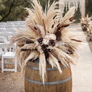 May include: A rustic wooden barrel is topped with a large floral arrangement. The arrangement features pampas grass, dried flowers, and burgundy roses. The barrel is held together with metal bands, and the scene is set outdoors.