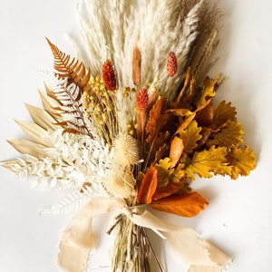 May include: A dried flower bouquet with a mix of white, beige, brown, orange, and red dried flowers. The bouquet is tied with a beige ribbon.