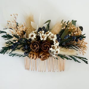 May include: A handcrafted hair comb featuring a natural arrangement of dried flowers, pinecones, and greenery. The comb includes white daisy-like flowers, dark blue berries, and tan grasses. The comb's teeth are a light gold color.