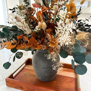 K&ouml;nnte beinhalten: Getrocknete Blumen in Braun-, Wei&szlig;- und Gr&uuml;nt&ouml;nen, die in einer grauen Keramikvase auf einem Holztablett mit goldenen Akzenten angeordnet sind.