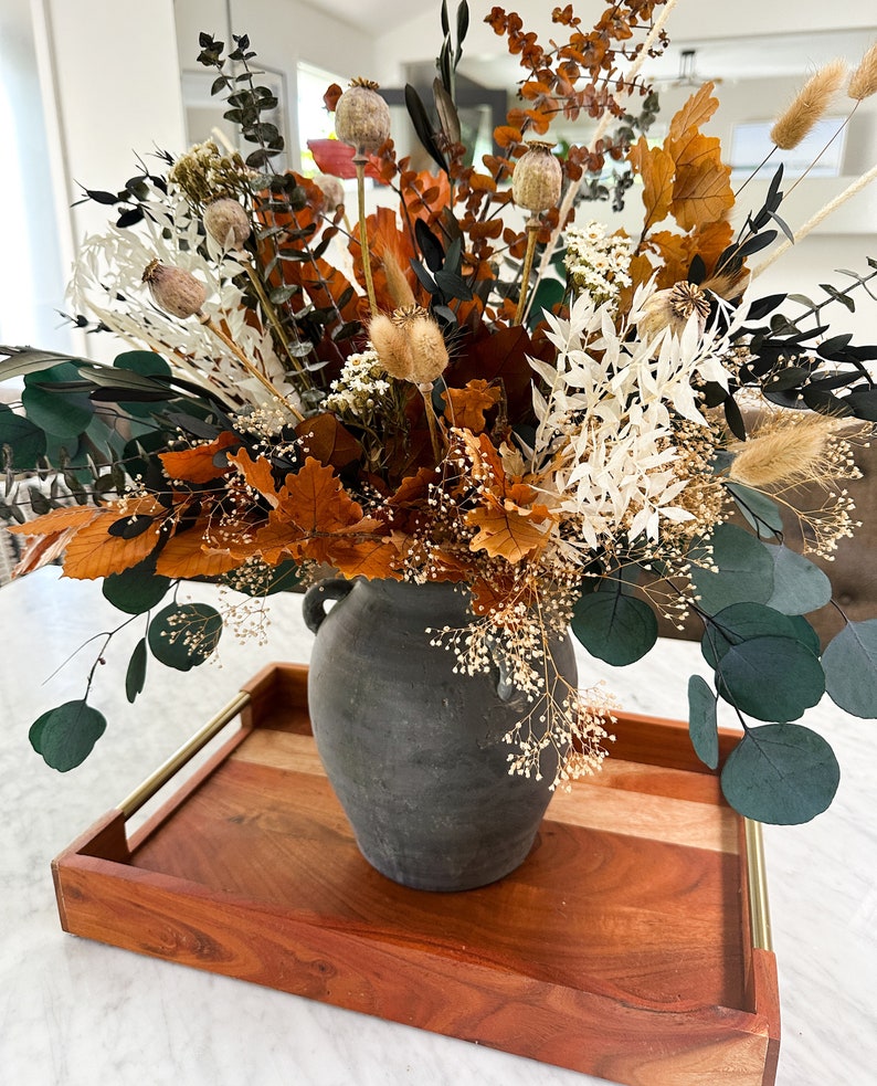 K&ouml;nnte beinhalten: Getrocknete Blumen in Braun-, Wei&szlig;- und Gr&uuml;nt&ouml;nen, die in einer grauen Keramikvase auf einem Holztablett mit goldenen Akzenten angeordnet sind.