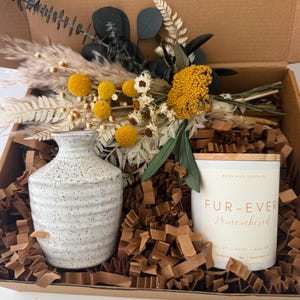 May include: A gift box containing a speckled white vase, a candle with a wooden lid, and a dried flower arrangement. The candle reads "FUR-EVER Remembered" with "Wildflowers + Sea Air" below. The box is filled with brown paper.