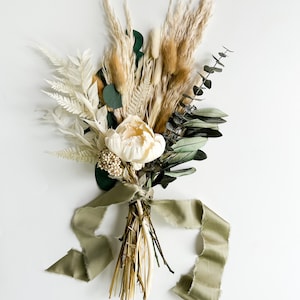 May include: A dried flower bouquet with a cream-colored peony, dried pampas grass, eucalyptus, and other dried foliage tied with a sage green ribbon.
