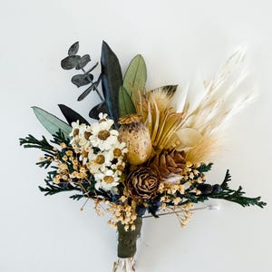 May include: A handcrafted boutonniere featuring a variety of dried flowers and foliage. The arrangement includes white daisies, dark green leaves, and tan pampas grass, bound with a dark green ribbon. A decorative pin secures the bouquet.
