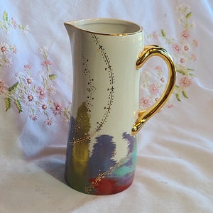 May include: A white ceramic pitcher with a gold handle and a colorful abstract design. The pitcher has a gold trim around the top and a gold design on the front.
