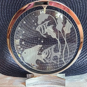 May include: A silver-toned metal stand with a round glass plate featuring an etched design of three fish and aquatic plants. The glass plate is surrounded by a silver-toned metal frame.