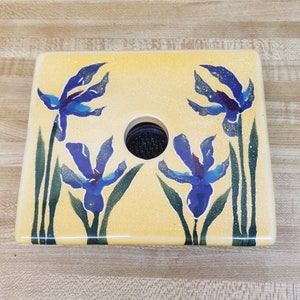 May include: A yellow ceramic flower frog with a blue and purple iris design. The frog has a round hole in the center for holding flowers.