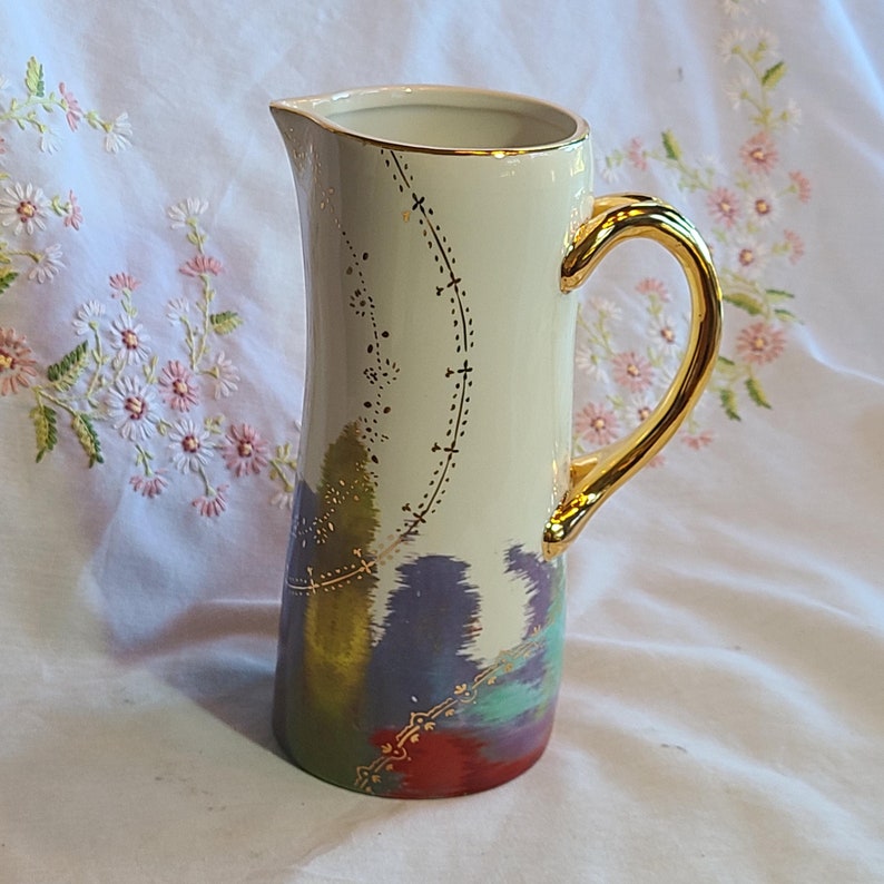 May include: A white ceramic pitcher with a gold handle and a colorful abstract design. The pitcher has a gold trim around the top and a gold design on the front.
