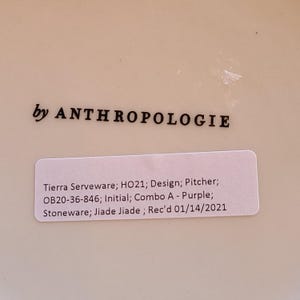 May include: A white ceramic pitcher with a sticker that reads "Tierra Serveware; HO21; Design; Pitcher; OB20-36-846; Initial; Combo A - Purple; Stoneware; Jiade Jiade; Rec'd 01/14/2021" and the text "by ANTHROPOLOGIE" printed on the bottom.