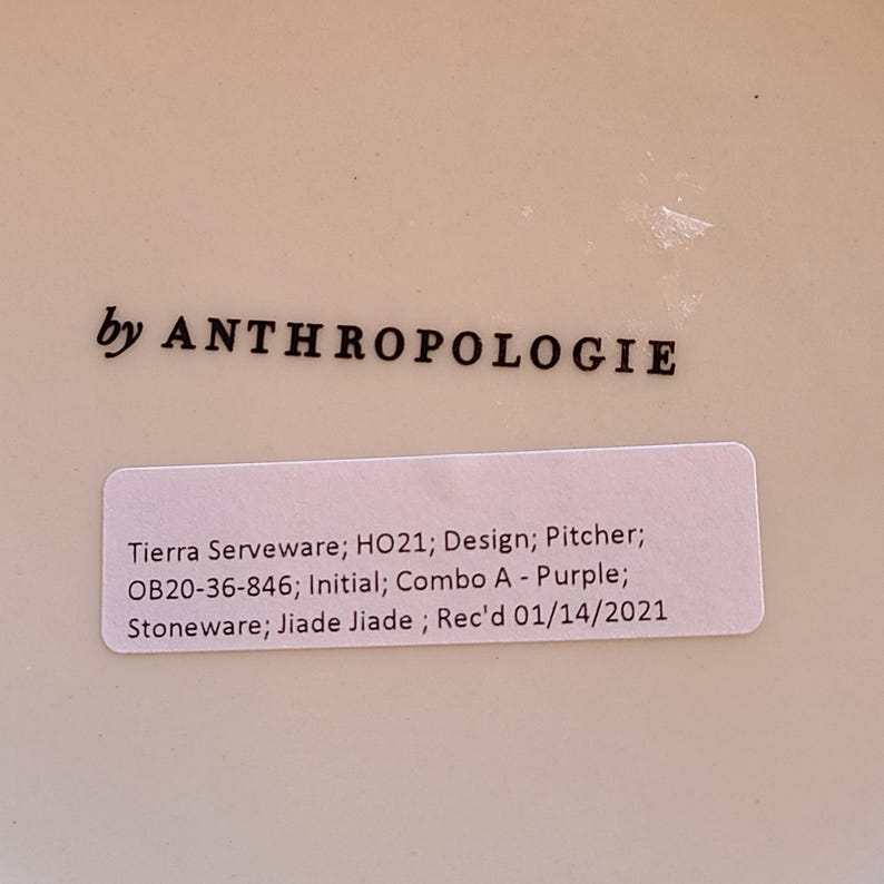 May include: A white ceramic pitcher with a sticker that reads "Tierra Serveware; HO21; Design; Pitcher; OB20-36-846; Initial; Combo A - Purple; Stoneware; Jiade Jiade; Rec'd 01/14/2021" and the text "by ANTHROPOLOGIE" printed on the bottom.