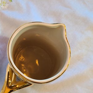 May include: A gold ceramic pitcher with a white interior. The pitcher has a wide mouth and a handle.