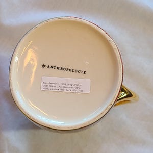May include: The bottom of a white ceramic pitcher with a gold handle. The pitcher has a red and gold design and the text "by ANTHROPOLOGIE" printed on the bottom. The text "Tierra Serveware; HO21; Design; Pitcher; 0820-36-846; Initial; Combo A-Purple; Stoneware; Jiade Jiade, Rec'd 01/14/2021" is printed on a white sticker.
