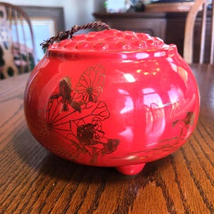 May include: A red ceramic jar with a lid and a brown rope handle. The jar features gold floral designs and rests on three small feet. The lid has a textured, dotted pattern.