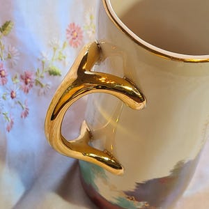 May include: A cream-colored ceramic pitcher with a gold-colored handle and rim. The handle is shaped like a branch and has a textured surface. The pitcher has a floral design on the side.