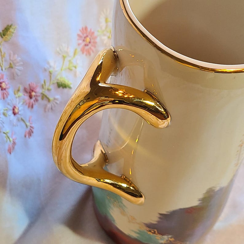 May include: A cream-colored ceramic pitcher with a gold-colored handle and rim. The handle is shaped like a branch and has a textured surface. The pitcher has a floral design on the side.