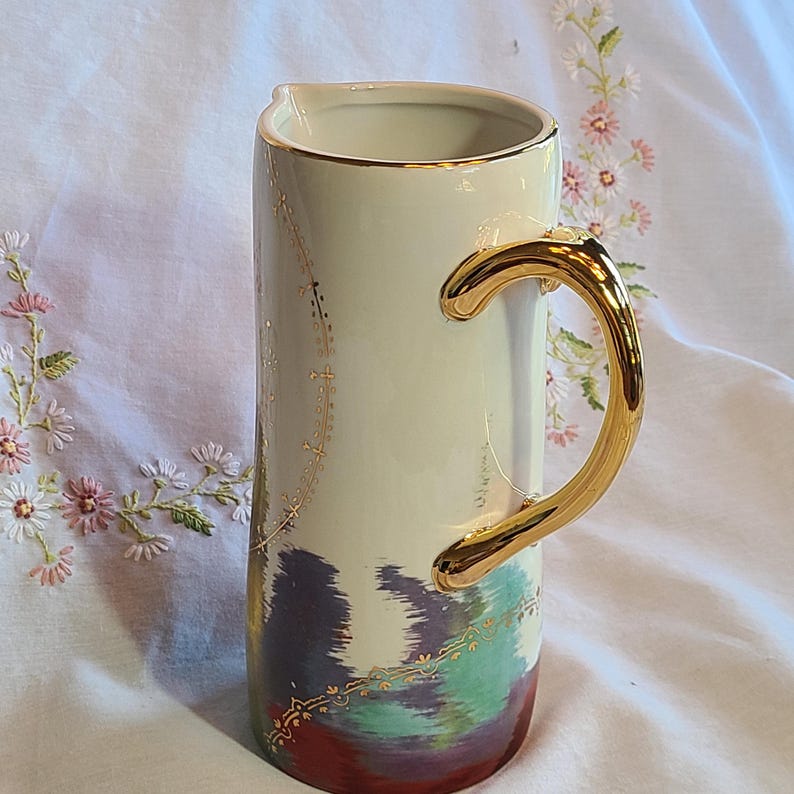 May include: A white ceramic pitcher with a gold handle and a colorful abstract design. The pitcher has a gold rim and a gold pattern around the top.