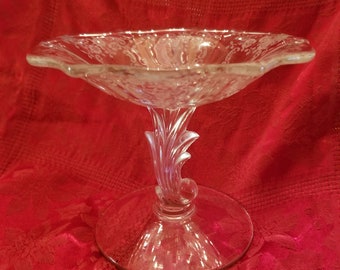 May include: A clear glass pedestal bowl with a scalloped edge and a swirl design. The bowl has a single, ornate stem with a scroll-like design.