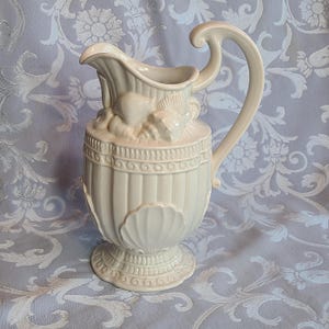 May include: A white ceramic pitcher with a seashell design. The pitcher has a wide base and a tall, slender body. The handle is curved and has a decorative design. The pitcher is decorated with seashells and other nautical motifs.