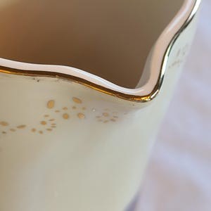 May include: Close-up of the rim of a white ceramic creamer with a gold trim and a delicate pattern of gold dots.