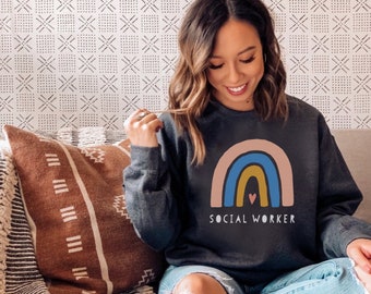 May include: A dark grey sweatshirt with a rainbow graphic and the text "SOCIAL WORKER". The rainbow is made up of pink, blue, yellow, and orange stripes. There is a small pink heart in the center of the rainbow.