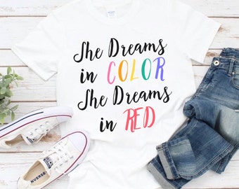 Puede incluir: Camiseta blanca con el texto "She Dreams in COLOR She Dreams in RED" en letras de colores.