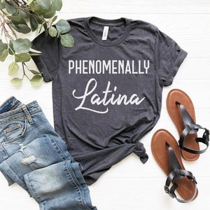 May include: Dark gray t-shirt with the words "PHENOMENALLY Latina" in white. The shirt is paired with blue jeans and brown sandals with black straps. The items are arranged on a white wooden surface.