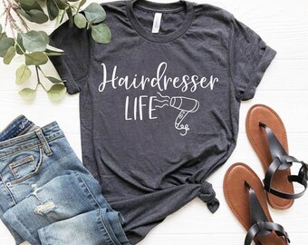 Può includere: T-shirt grigio scuro con le parole "Hairdresser LIFE" e un disegno a tratto di un asciugacapelli. La maglietta è abbinata a jeans blu e sandali marroni con cinturini neri. Un rametto di verde è sullo sfondo.