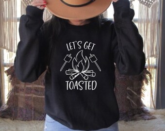 Puede incluir: Sudadera negra con texto blanco que dice "Let's Get Toasted" y un gráfico de una fogata con malvaviscos en palos.