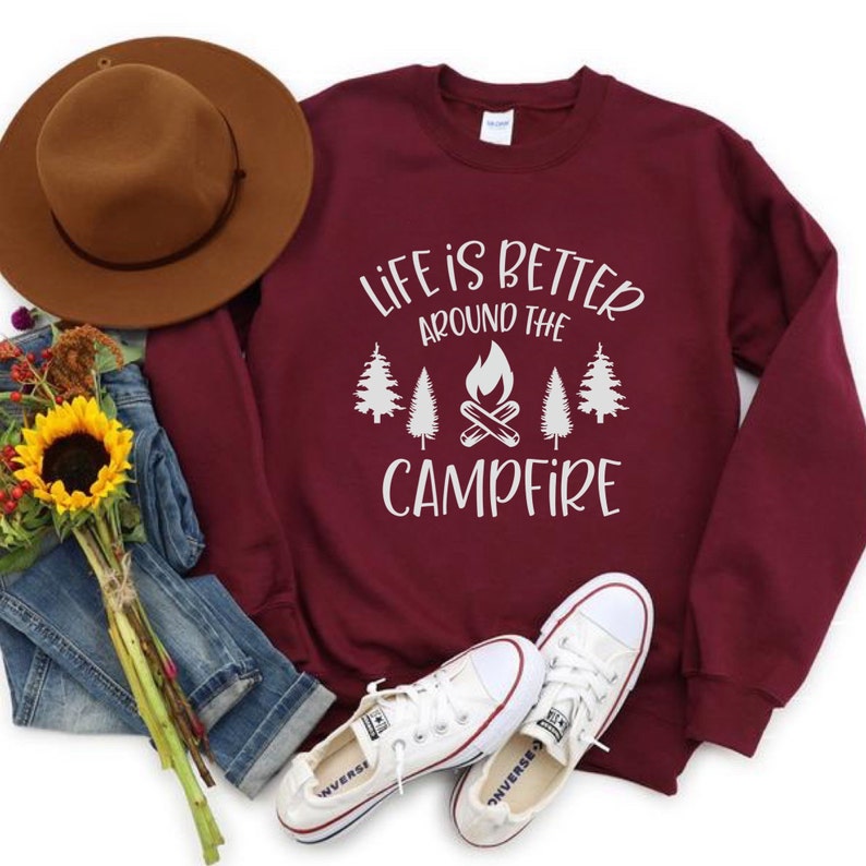Puede incluir: Sudadera color burdeos con texto blanco que dice "Life is better around the campfire" con un gr&aacute;fico de una fogata y &aacute;rboles.