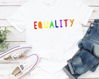May include: White t-shirt with the word "EQUALITY" printed in rainbow colors. The shirt is laid flat on a wooden surface with a pair of white sneakers and blue jeans.