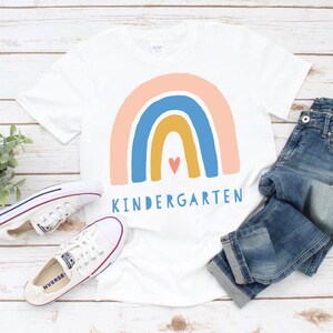 Puede incluir: Camiseta blanca con un dise&ntilde;o de arco&iacute;ris y la palabra "KINDERGARTEN" impresa en ella. El arco&iacute;ris est&aacute; hecho de colores rosa, azul, amarillo y naranja. Hay un peque&ntilde;o coraz&oacute;n rojo en el centro del arco&iacute;ris.