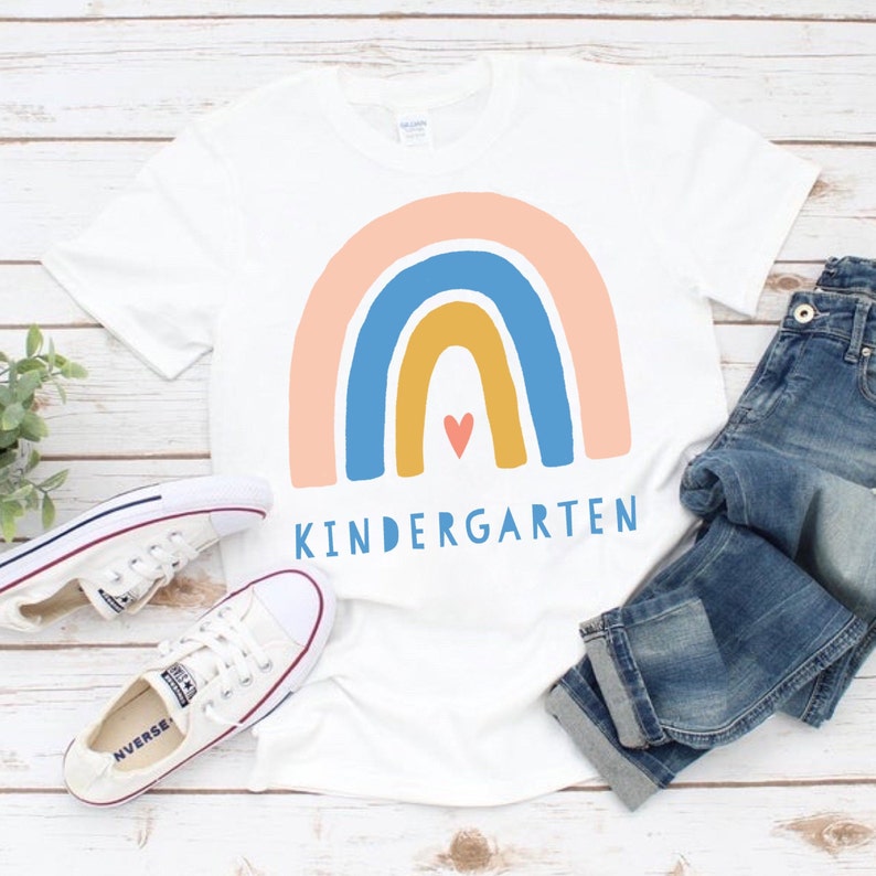 Puede incluir: Camiseta blanca con un dise&ntilde;o de arco&iacute;ris y la palabra "KINDERGARTEN" impresa en ella. El arco&iacute;ris est&aacute; hecho de colores rosa, azul, amarillo y naranja. Hay un peque&ntilde;o coraz&oacute;n rojo en el centro del arco&iacute;ris.