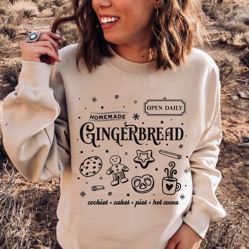 Może przedstawiać: Beżowa bluza z czarnym nadrukiem graficznym, na kt&oacute;rym widnieje napis "Homemade Gingerbread" wraz z piernikowym ludzikiem, ciasteczkami, preclami i filiżanką gorącej czekolady. Tekst pod grafiką brzmi "cookies - cakes - pies - hot cocoa".