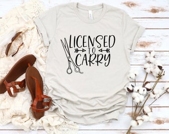 以下が含まれることがあります： 「Licensed to Carry」という文字とハサミがプリントされた薄いグレーのTシャツ