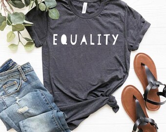 May include: A dark gray t-shirt with the word "EQUALITY" printed in white on the front. The shirt is folded and laid on a white surface with a pair of brown sandals and blue jeans.